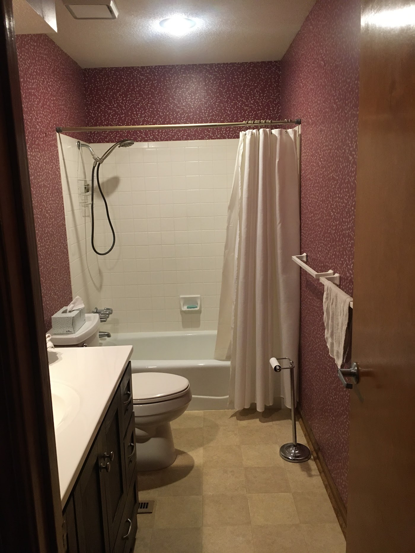 Bathroom before remodel with burgundy patterned wallpaper, dated square tile, and a tub-shower